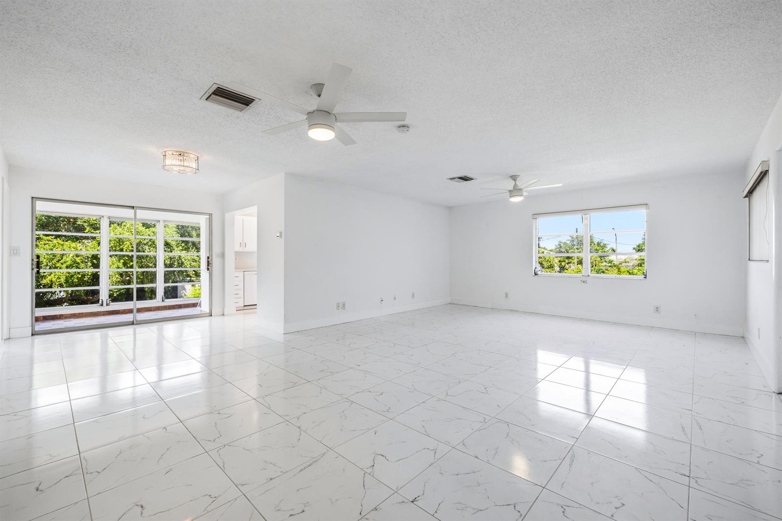 A bright, empty living room features white marble-look tile floors, two ceiling fans, and sliding doors — 941 Crystal Lake Drive # 207, Deerfield Beach FL 33064, DEERFIELD BEACH, FL