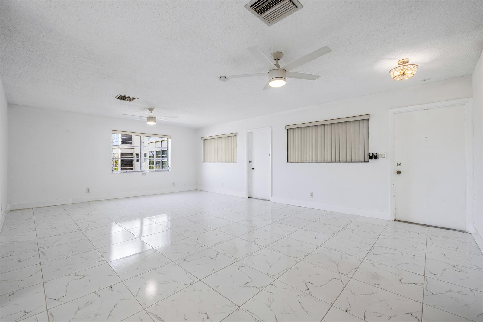 A bright, empty living room features white marble-look tile flooring, two ceiling fans, and windows — 941 Crystal Lake Drive # 207, Deerfield Beach FL 33064, DEERFIELD BEACH, FL