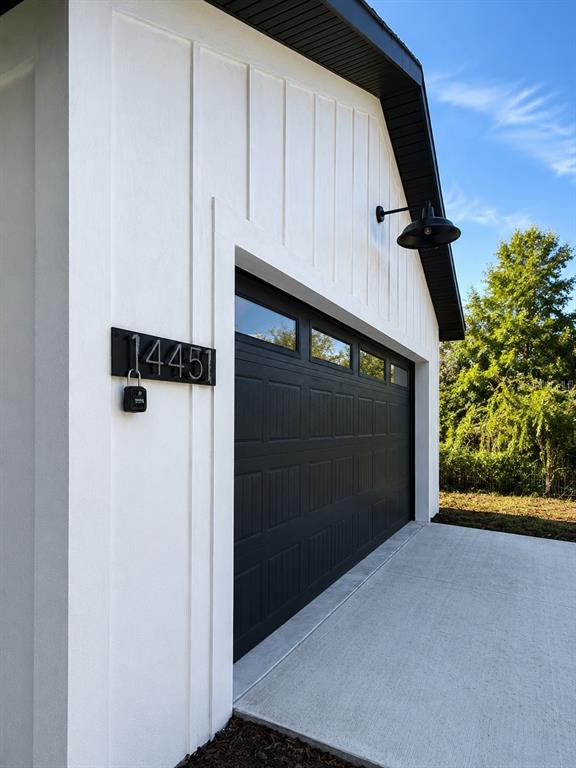 A white board-and-batten garage exterior features a black door, a gooseneck light, and house numbers — 14451 SE 43RD TERRACE, SUMMERFIELD, FL