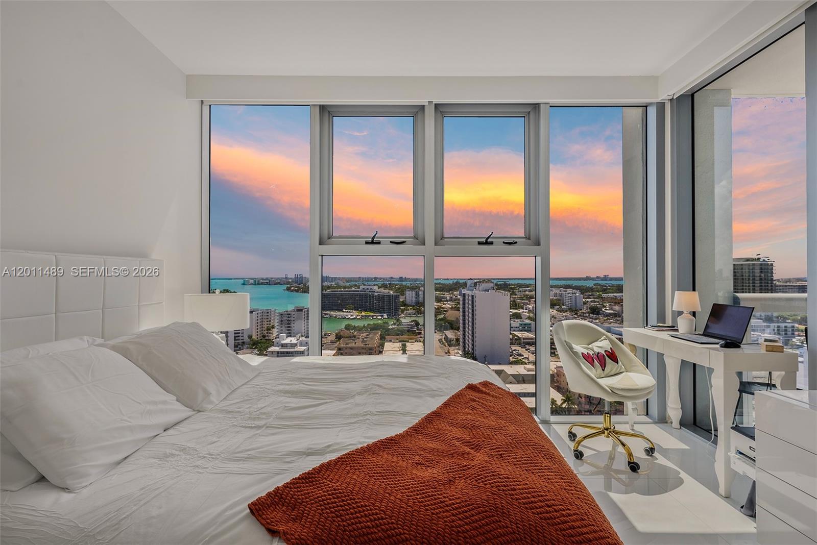 A bedroom features a white bed, a desk, and floor-to-ceiling windows overlooking a city skyline — 6899 Collins Ave # 2004, Miami Beach FL 33141, MIAMI BEACH, FL