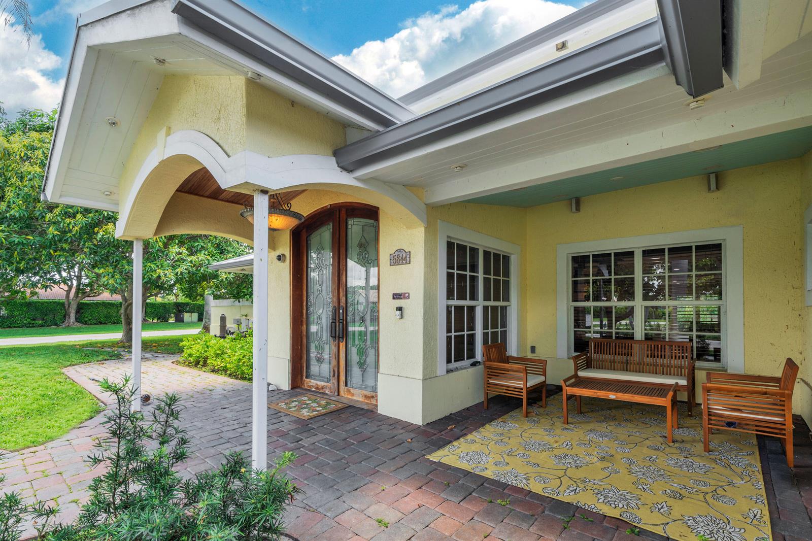 A yellow house exterior features a covered brick patio with wooden furniture and double doors — 5844 Pinebrook Drive, Boca Raton FL 33433, BOCA RATON, FL