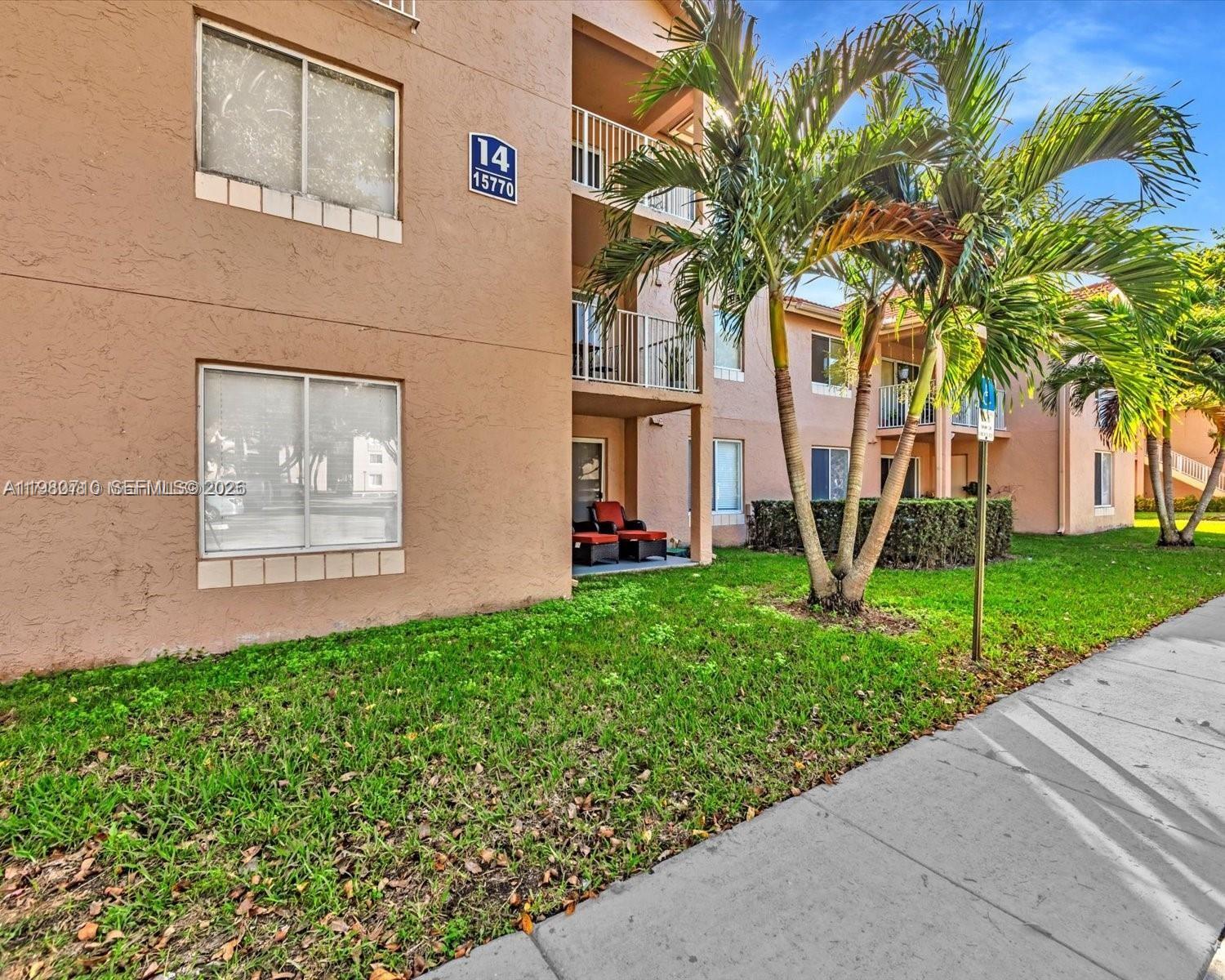 An exterior view of a multi-story tan apartment building with palm trees and a sidewalk — 15770 SW 106th Ter # 303, Miami FL 33196, MIAMI, FL