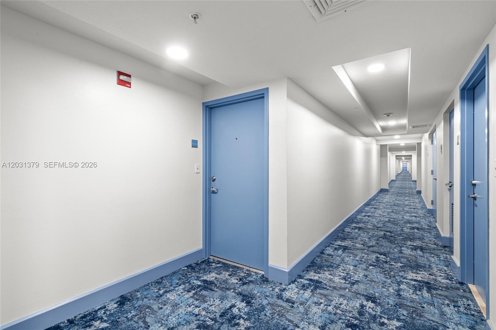 A long apartment hallway features blue doors, white walls, and patterned blue carpeting — 7600 Collins Ave # 1210, Miami Beach FL 33141, MIAMI BEACH, FL