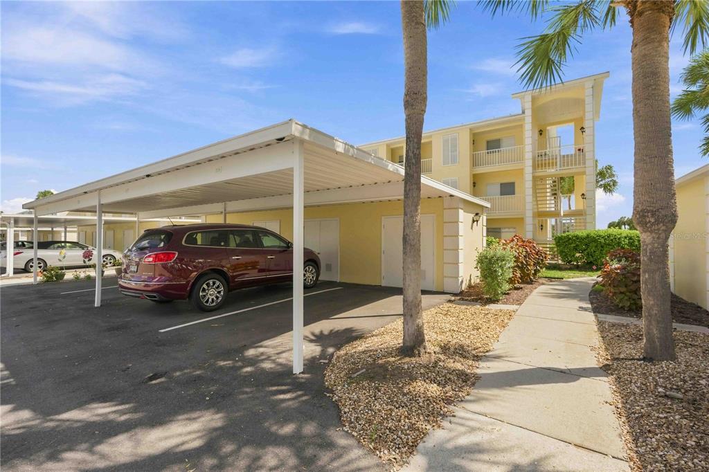 A covered parking carport sits next to a multi-story yellow apartment building with balconies — 406 CERROMAR CIRCLE N UNIT 328, VENICE, FL