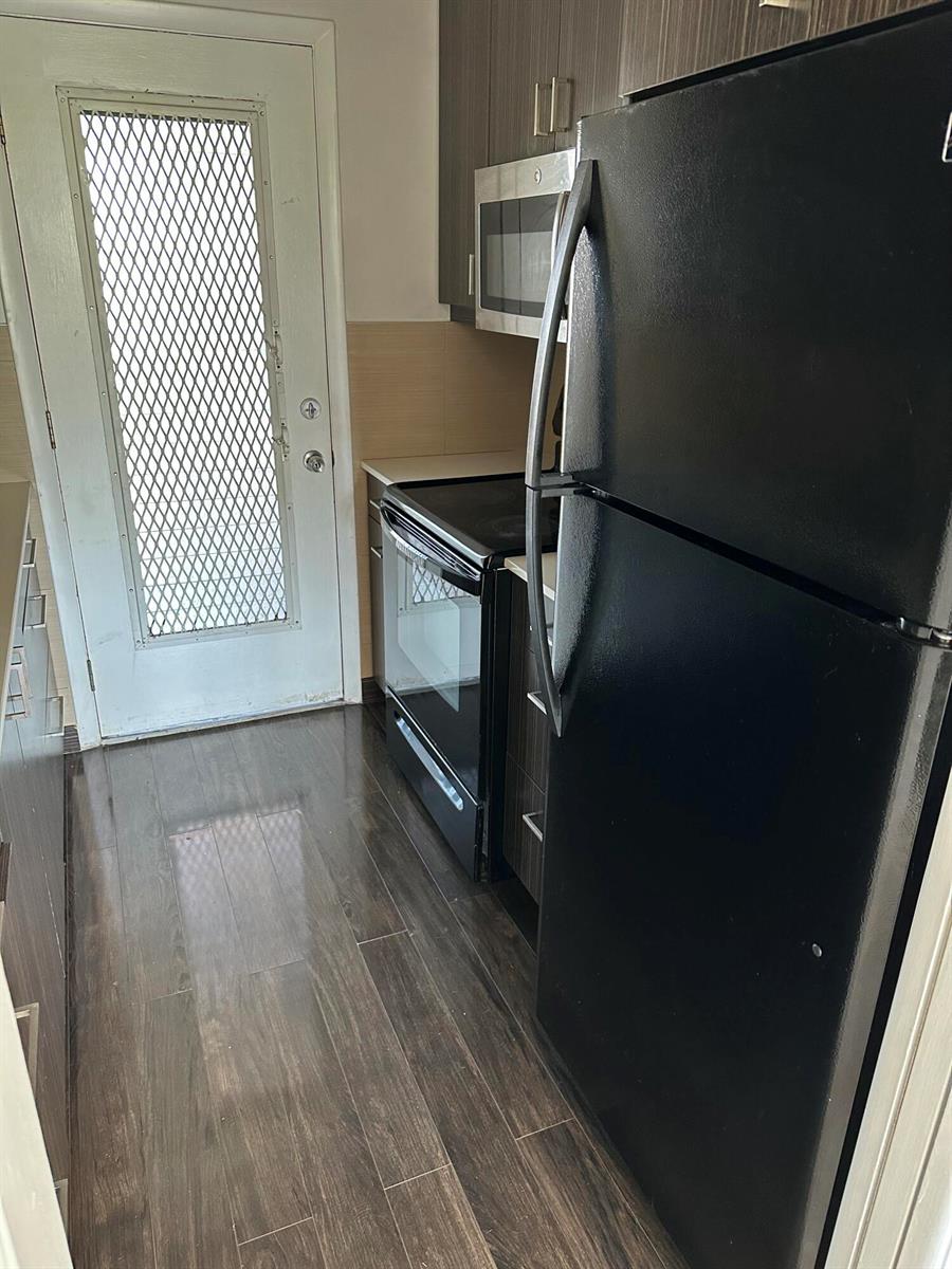 A narrow kitchen features dark wood-look flooring, dark cabinetry, a black refrigerator, and a glass-paneled door — 1480 E Sample Road # 207, Pompano Beach FL 33064, POMPANO BEACH, FL