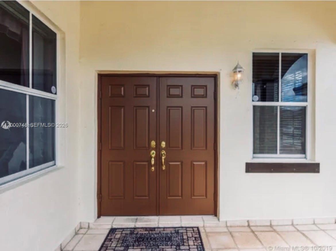A brown double front door is centered between two windows on a tiled porch — 8135 NW 198th Ter, Hialeah FL 33015, HIALEAH, FL