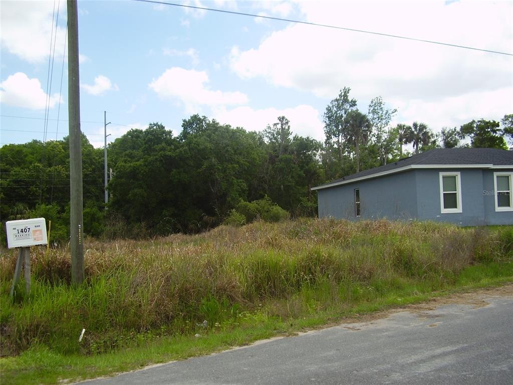 A vacant, grass-covered lot sits next to a gray house under a cloudy sky — 1409 TEAL DRIVE, KISSIMMEE, FL