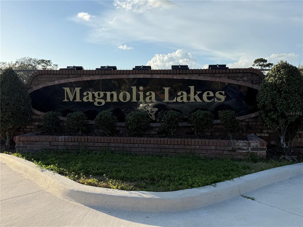 A brick entrance sign for Magnolia Lakes sits behind a curved concrete curb and landscaping — 3916 MAGNOLIA LAKE LANE, ORLANDO, FL