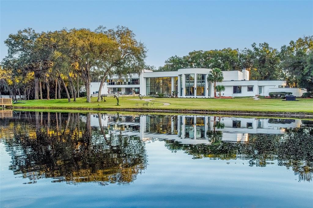A modern white house with large glass windows sits beside a calm lake with trees — 812 SWEETWATER CLUB BOULEVARD, LONGWOOD, FL