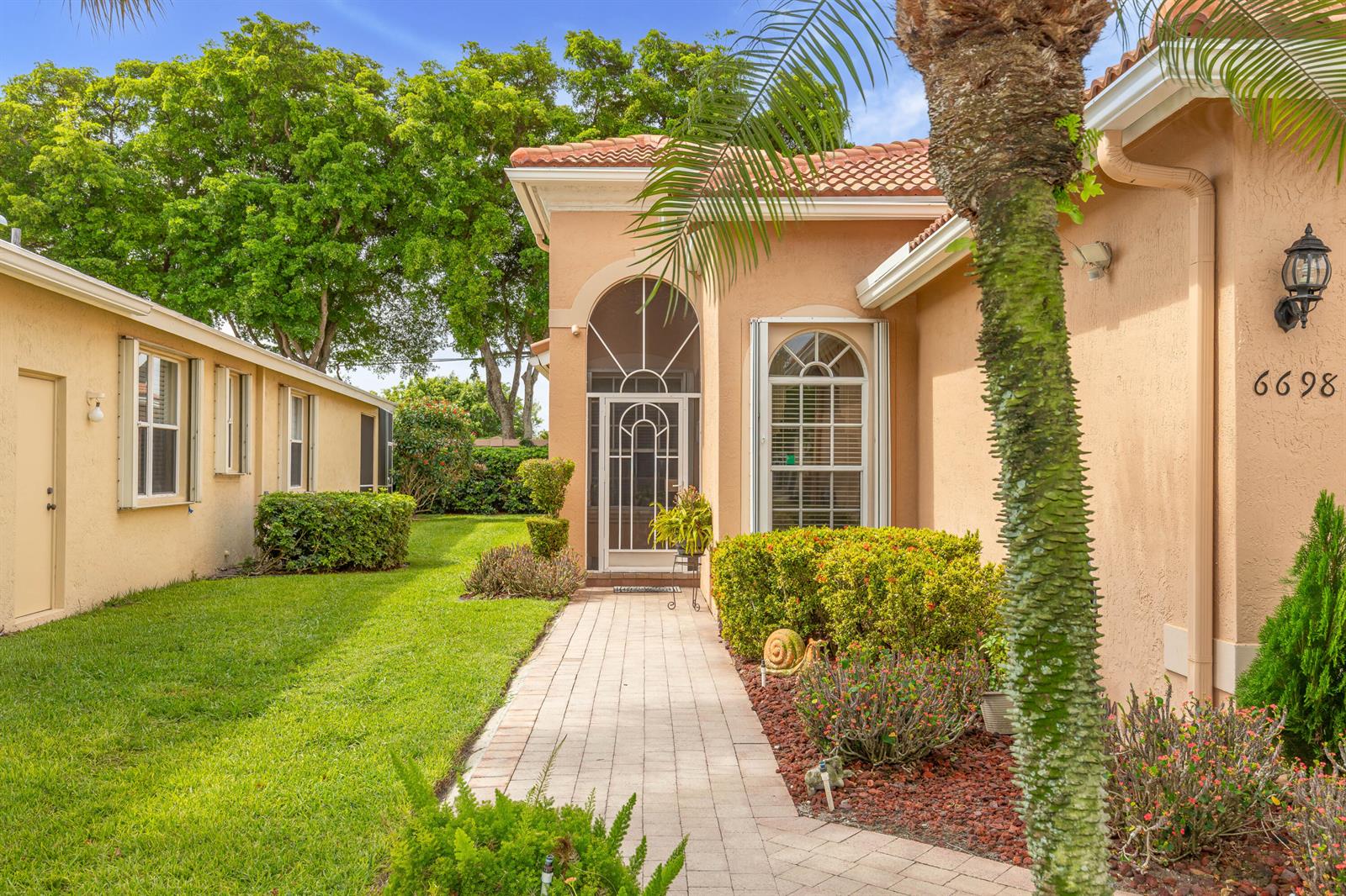 A paved walkway leads to the front entrance of a tan house with arched windows — 6698 Vía Roma, Delray Beach FL 33446, DELRAY BEACH, FL