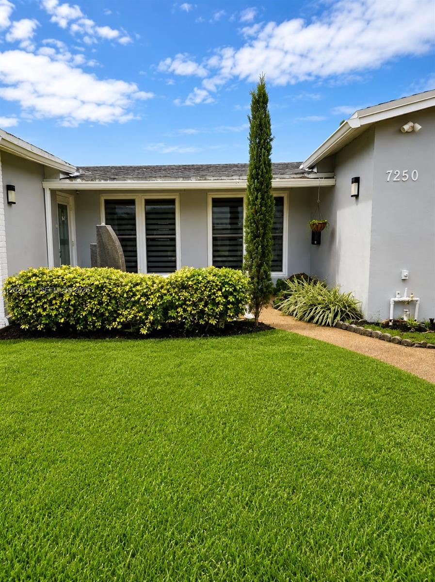 A single-story house exterior features a green lawn, a tall cypress tree, and landscaping — 7250 Dilido Blvd, Miramar FL 33023, MIRAMAR, FL
