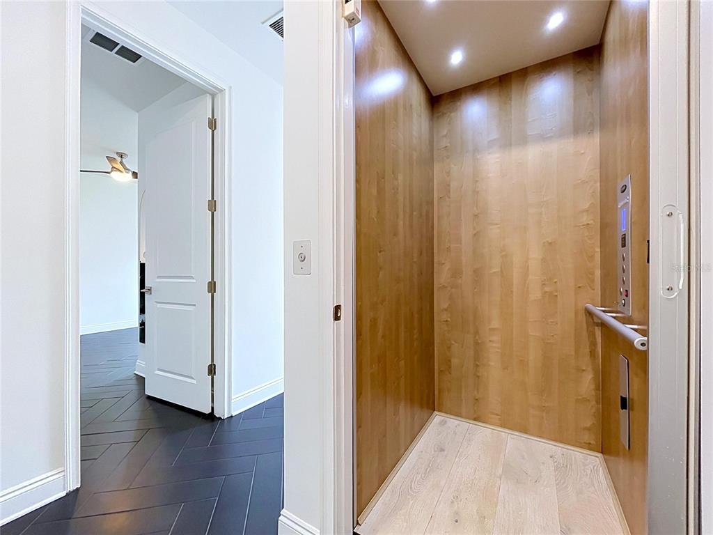 A residential elevator with wood-paneled walls sits next to a room with dark herringbone flooring — 3702 W ROLAND STREET UNIT 3, TAMPA, FL
