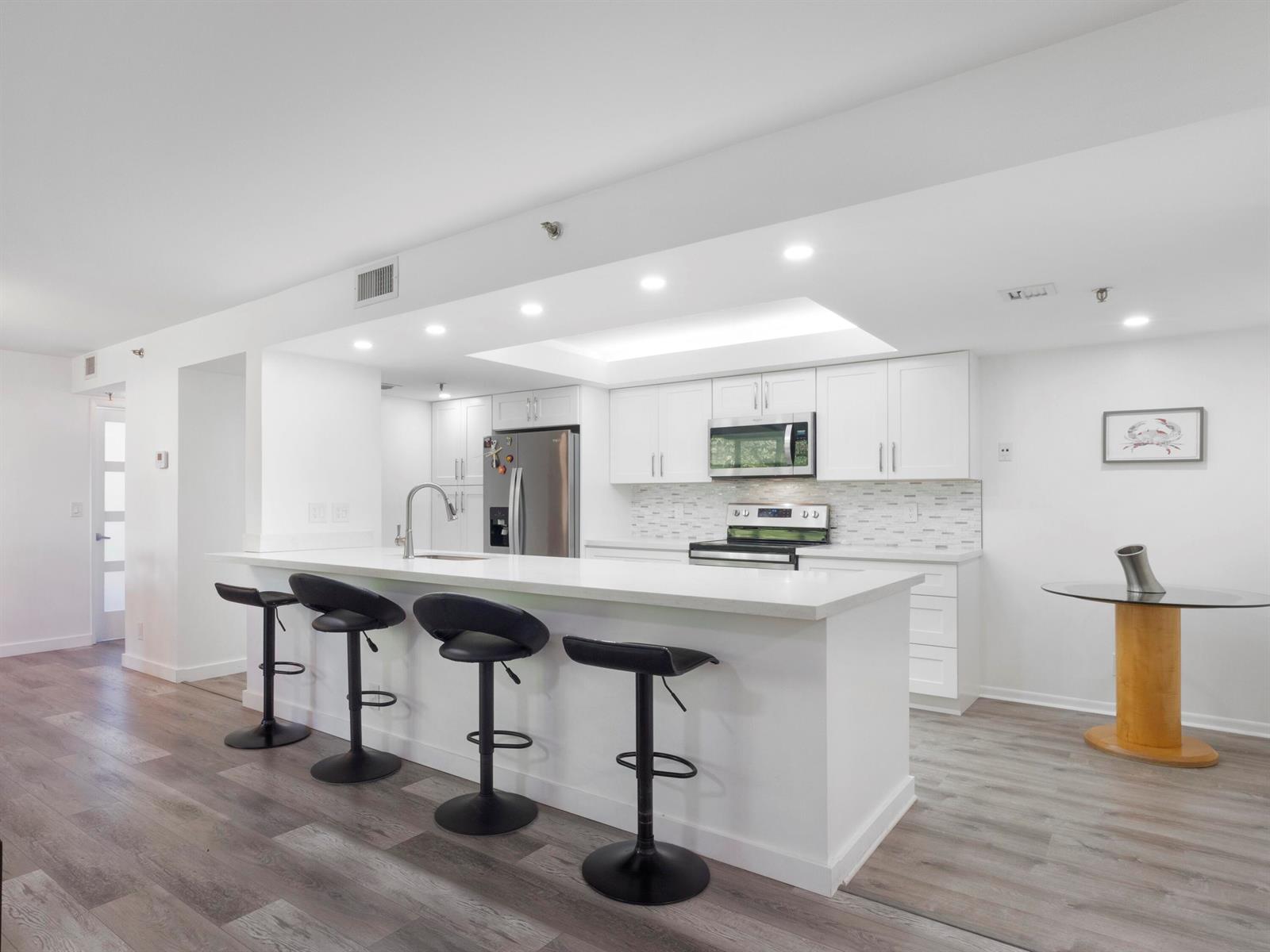 A modern kitchen features white cabinetry, a quartz breakfast bar with stools, and wood flooring — 955 Egret Circle # 402, Delray Beach FL 33444, DELRAY BEACH, FL