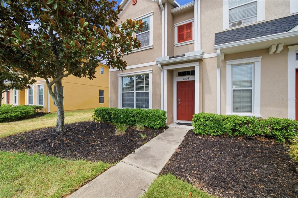 A tan two-story townhouse features a red front door, white trim, and landscaped front yard — 6924 TOWERING SPRUCE DRIVE, RIVERVIEW, FL