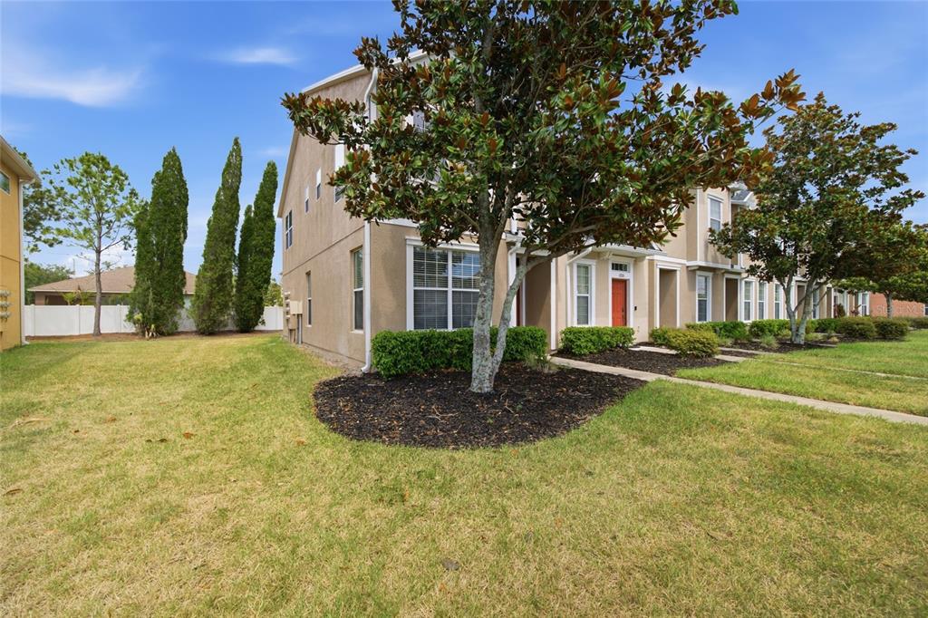 A two-story townhouse exterior with a tan stucco facade, a front tree, and grass lawn — 6924 TOWERING SPRUCE DRIVE, RIVERVIEW, FL