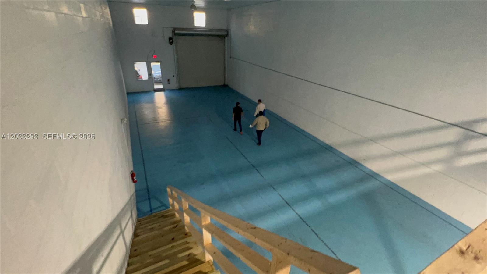 A high-ceiling warehouse space features a blue floor, a roll-up door, and wooden stairs — 1118 25th St # 3, West Palm Beach FL 33407, WEST PALM BEACH, FL