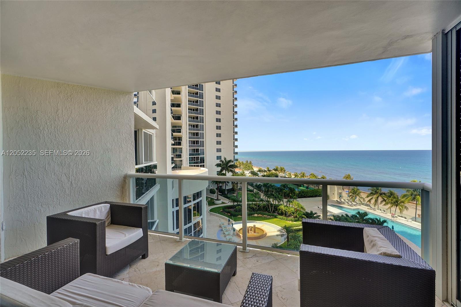 A balcony with wicker furniture overlooks a swimming pool, landscaped grounds, and the ocean — 19111 Collins Ave # 607, Sunny Isles Beach FL 33160, SUNNY ISLES BEACH, FL