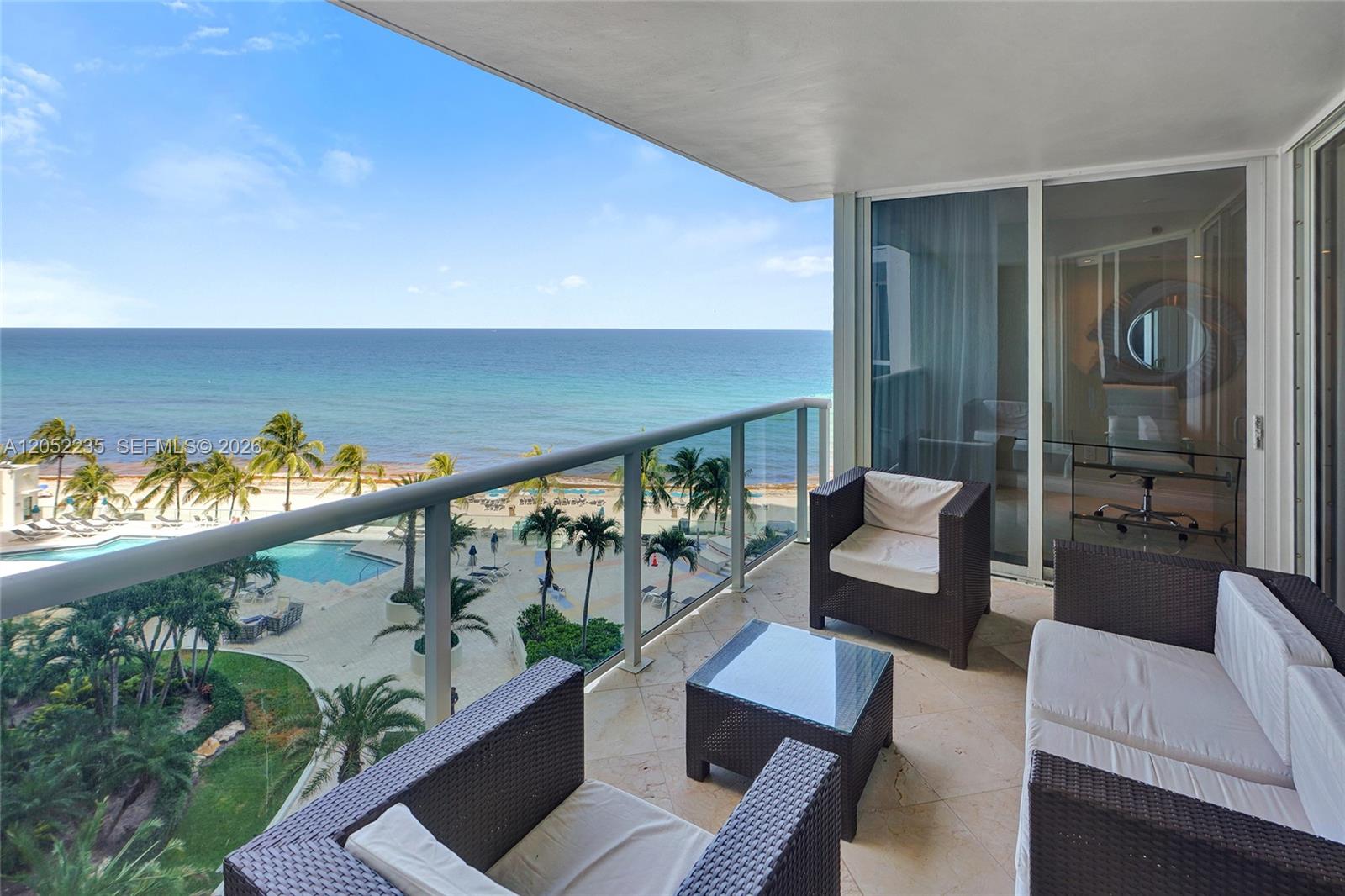 A balcony with wicker furniture overlooks a swimming pool, palm trees, and the ocean — 19111 Collins Ave # 607, Sunny Isles Beach FL 33160, SUNNY ISLES BEACH, FL