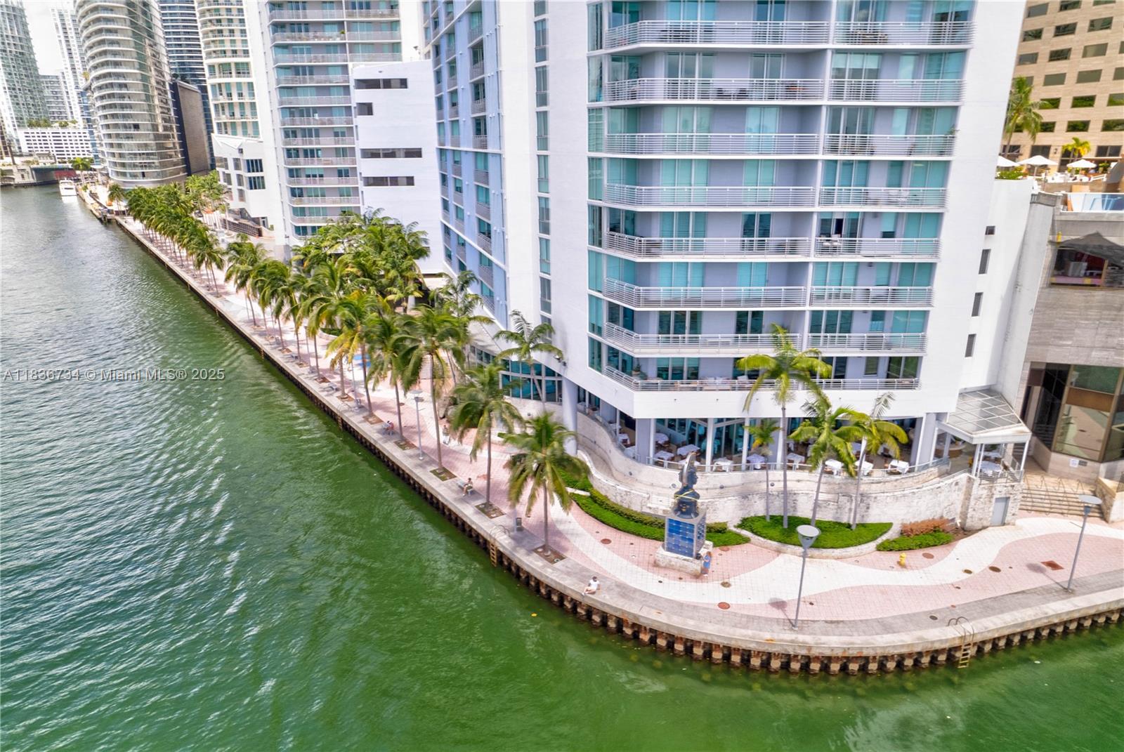 An aerial view shows a waterfront promenade with palm trees beside a modern high-rise building — 335 S Biscayne Blvd # 3108, Miami FL 33131, MIAMI, FL
