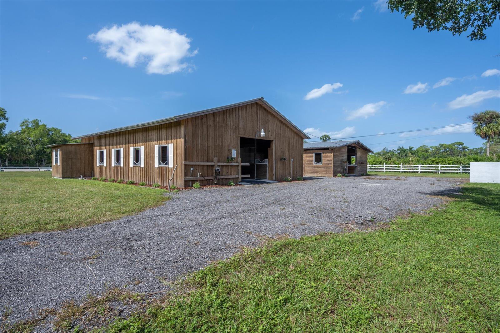A gravel driveway leads to two wooden horse barns under a bright blue sky — 13717 E Citrus Drive, FL 33470, FL