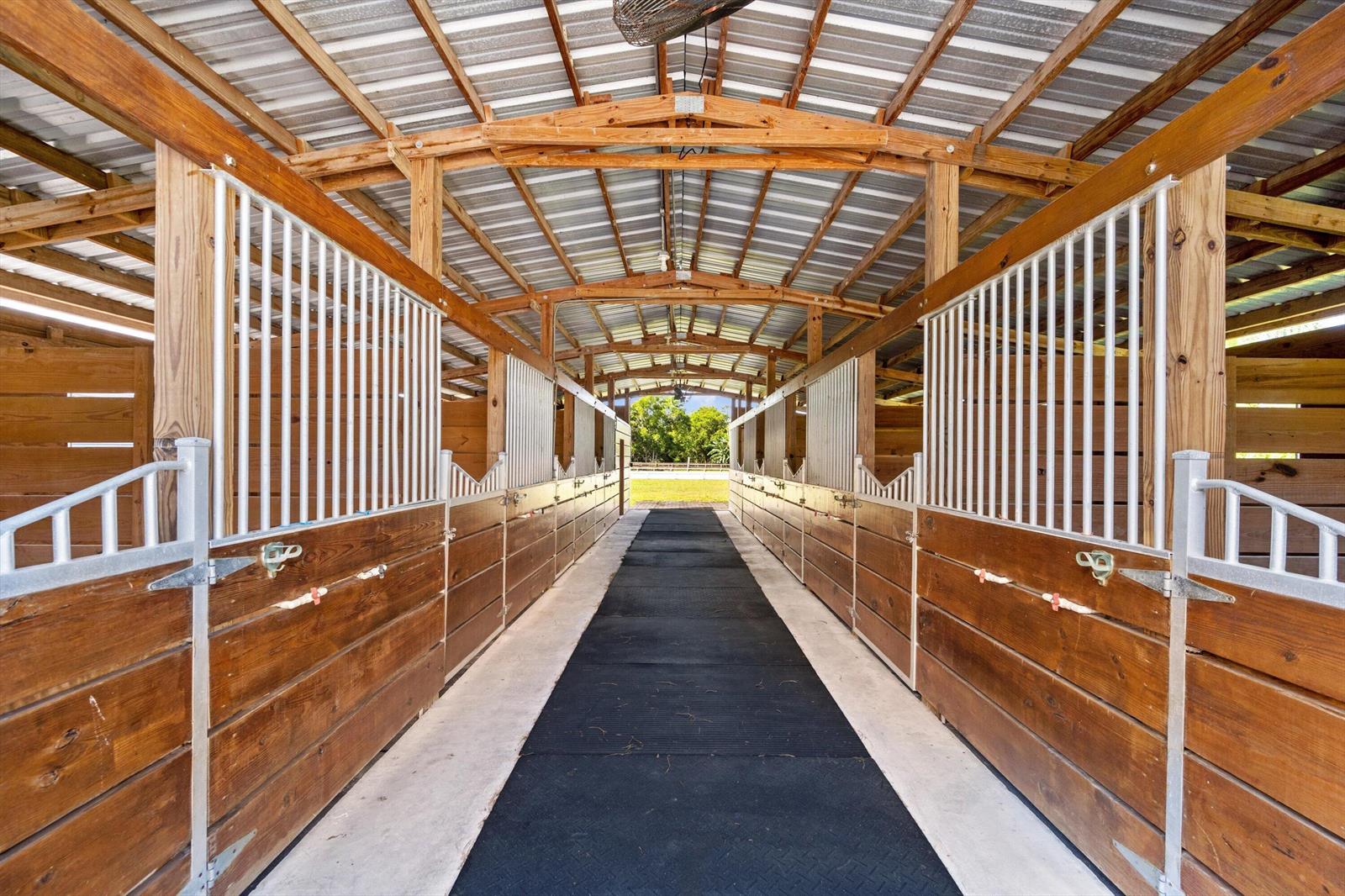 A horse barn aisle features rows of wooden stalls with metal grilles and rubber flooring — 13717 E Citrus Drive, FL 33470, FL