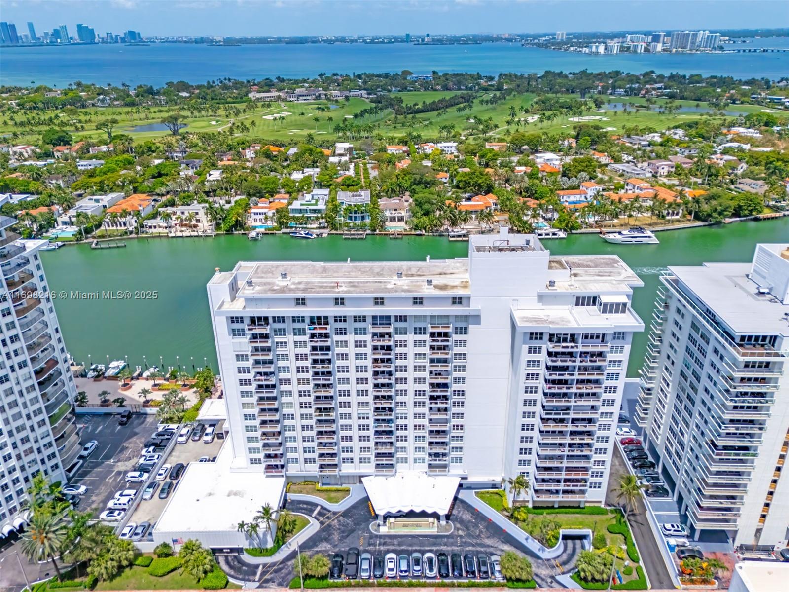 An aerial view shows a white high-rise apartment building next to a canal and golf course — 5700 Collins Ave # 5B, Miami Beach FL 33140, MIAMI BEACH, FL