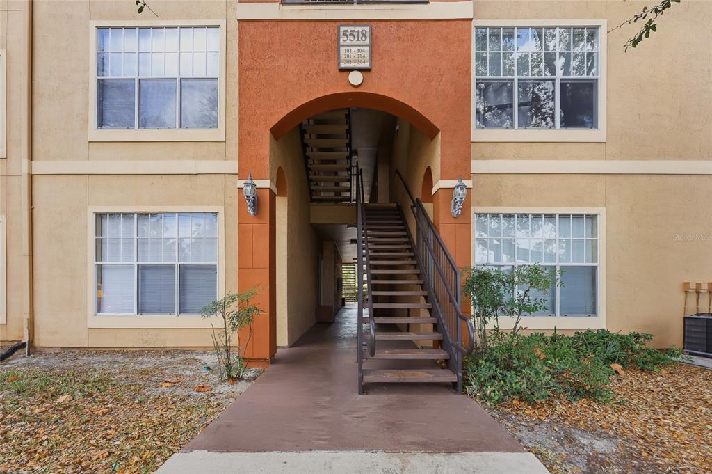 An exterior view of a three-story apartment building featuring a central staircase and arched entryway — 5518 METROWEST BOULEVARD UNIT 11-104, ORLANDO, FL