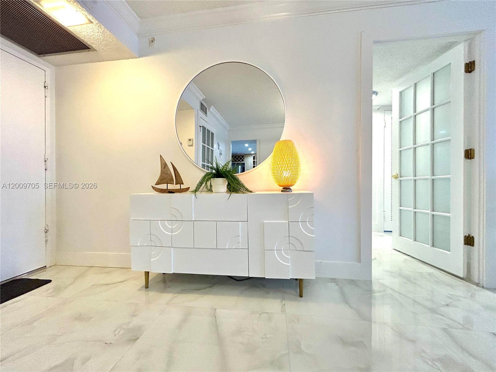 A white entryway features a modern sideboard, a round mirror, and marble-look tile flooring — 6039 Collins Ave # 1507, Miami Beach FL 33140, MIAMI BEACH, FL