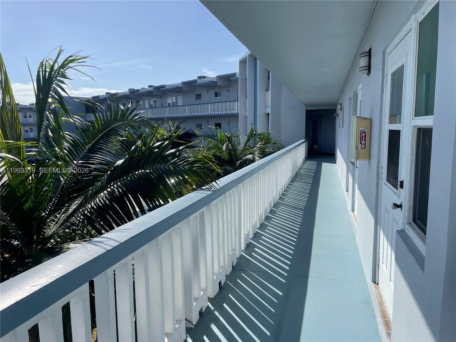 An outdoor walkway with a white railing overlooks palm trees and a neighboring building — 2300 Park Ln # 208, Hollywood FL 33021, HOLLYWOOD, FL