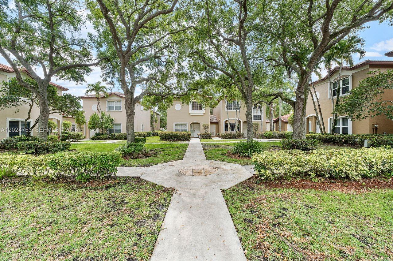 A concrete walkway leads through a grassy courtyard toward multi-story residential buildings with trees — 16035 Emerald Cove Rd, Weston FL 33331, WESTON, FL
