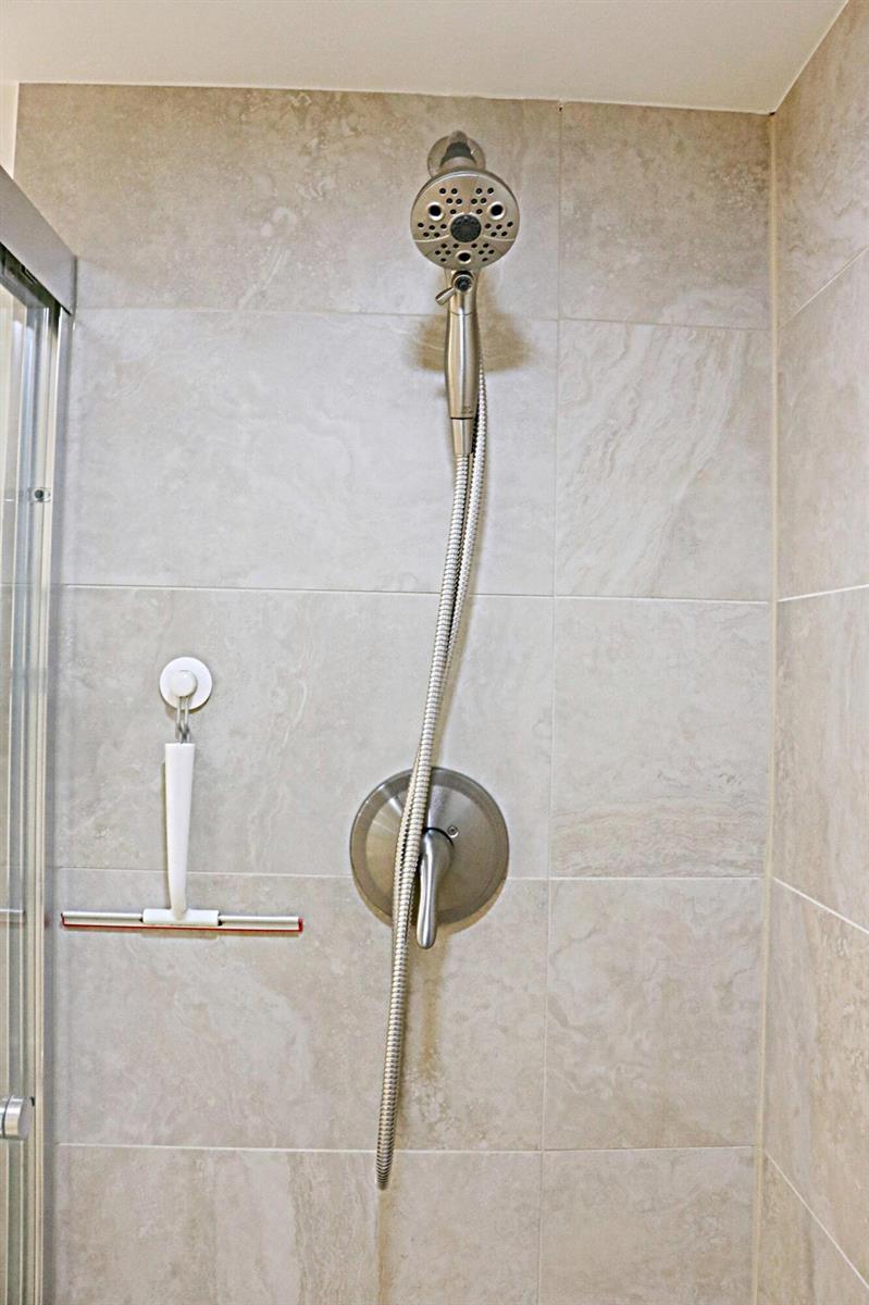 A shower stall features beige stone-look wall tiles, a handheld showerhead, and a wall-mounted squeegee — 230 NE 26th Avenue # 1070, Boynton Beach FL 33435, BOYNTON BEACH, FL