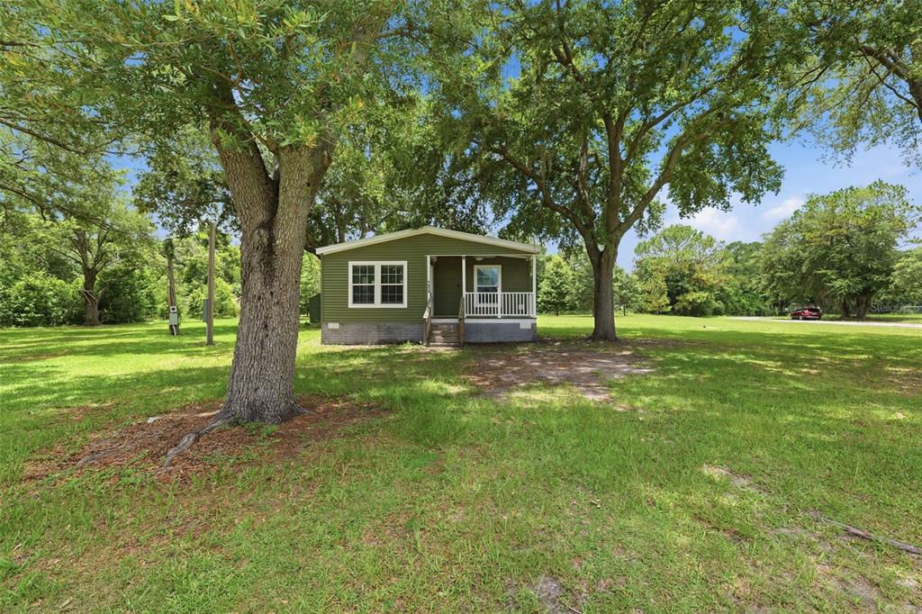 A green single-story house with a front porch sits on a large, grassy, tree-filled lot — 7239 ROOKS DRIVE, WESLEY CHAPEL, FL