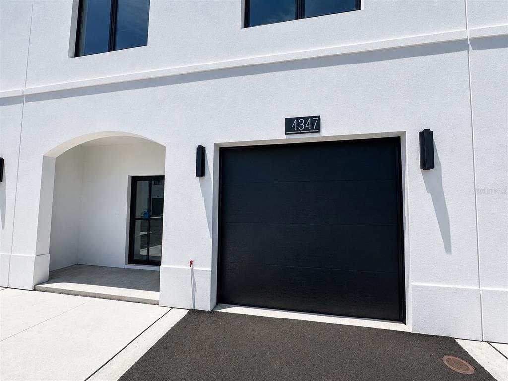 A white exterior wall features a black garage door, a recessed entryway, and modern lighting — 4347 CAMBIO GARDENS COURT, TAMPA, FL
