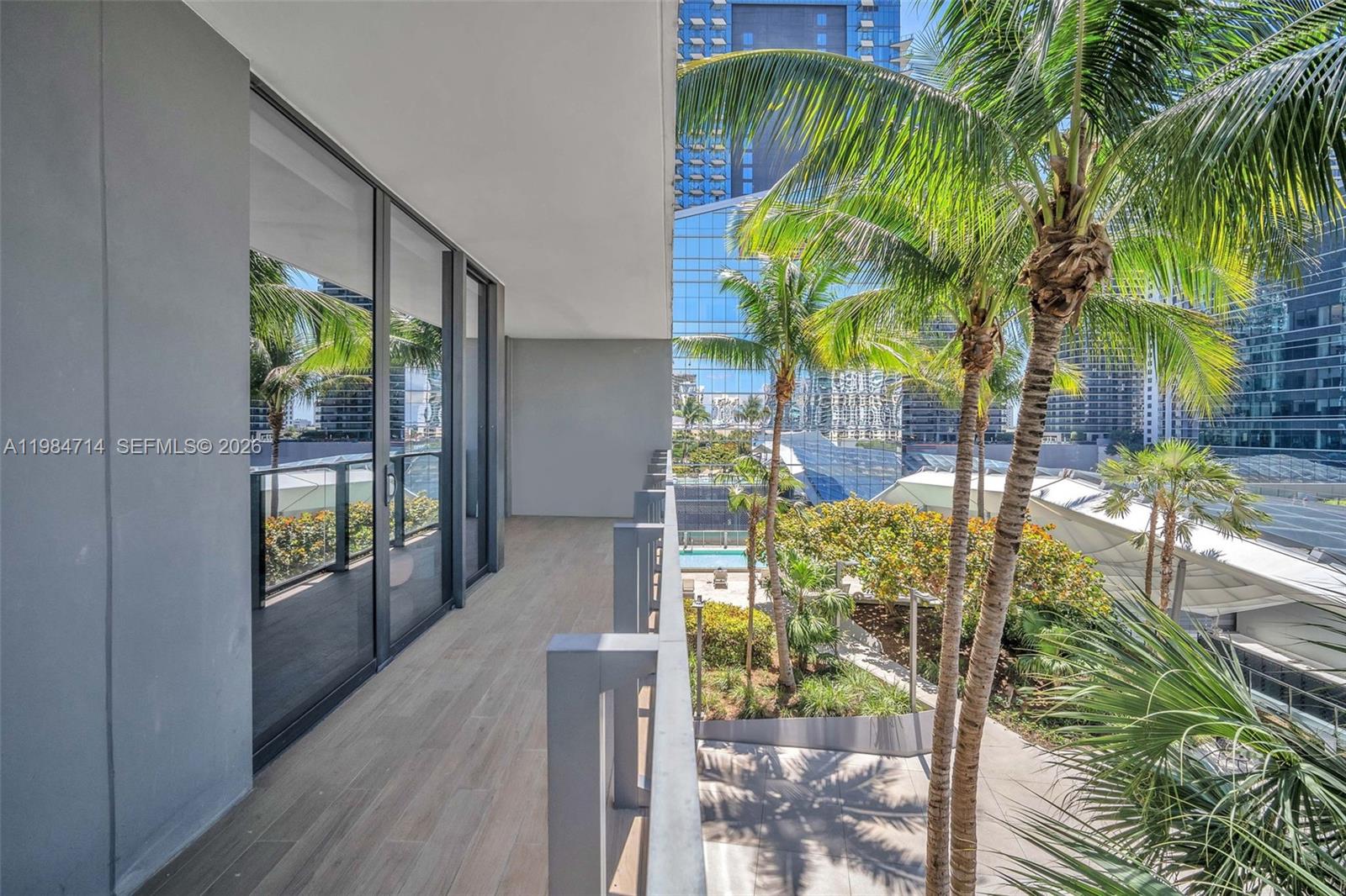 A modern balcony features wood-look tile flooring, glass railings, and views of palm trees — 68 SE 6th St # 806, Miami FL 33131, MIAMI, FL