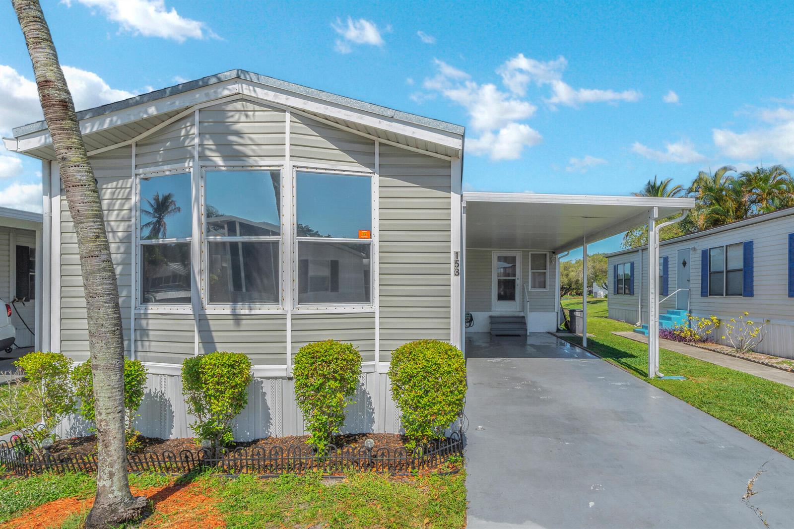 A light gray manufactured home features a front carport, large windows, and manicured landscaping — 153 Plantation Boulevard, Lake Worth FL 33467, LAKE WORTH, FL