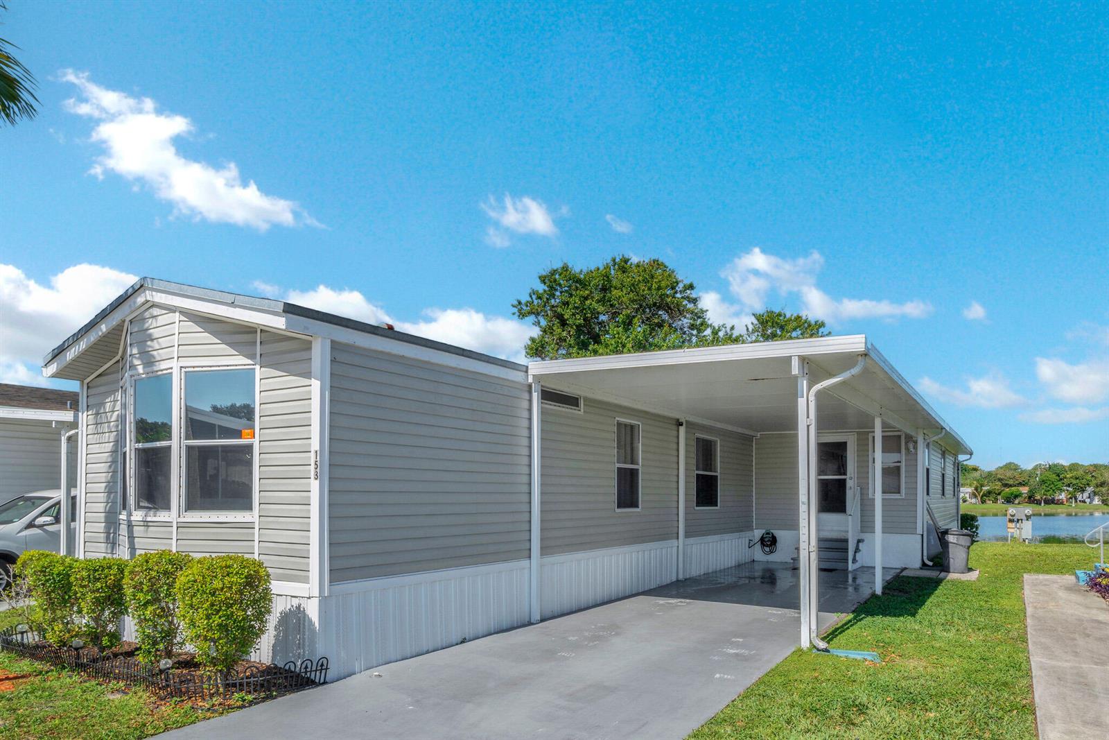 A light gray manufactured home features a covered carport and a small front yard — 153 Plantation Boulevard, Lake Worth FL 33467, LAKE WORTH, FL