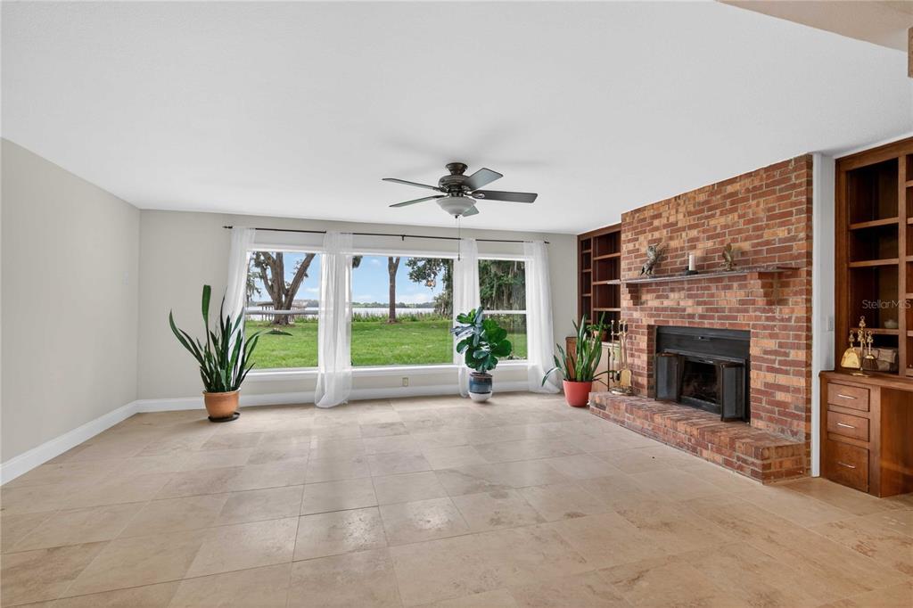 A living room features a brick fireplace, tile flooring, and large windows overlooking a lake — 5616 TRIMBLE PARK ROAD, MOUNT DORA, FL