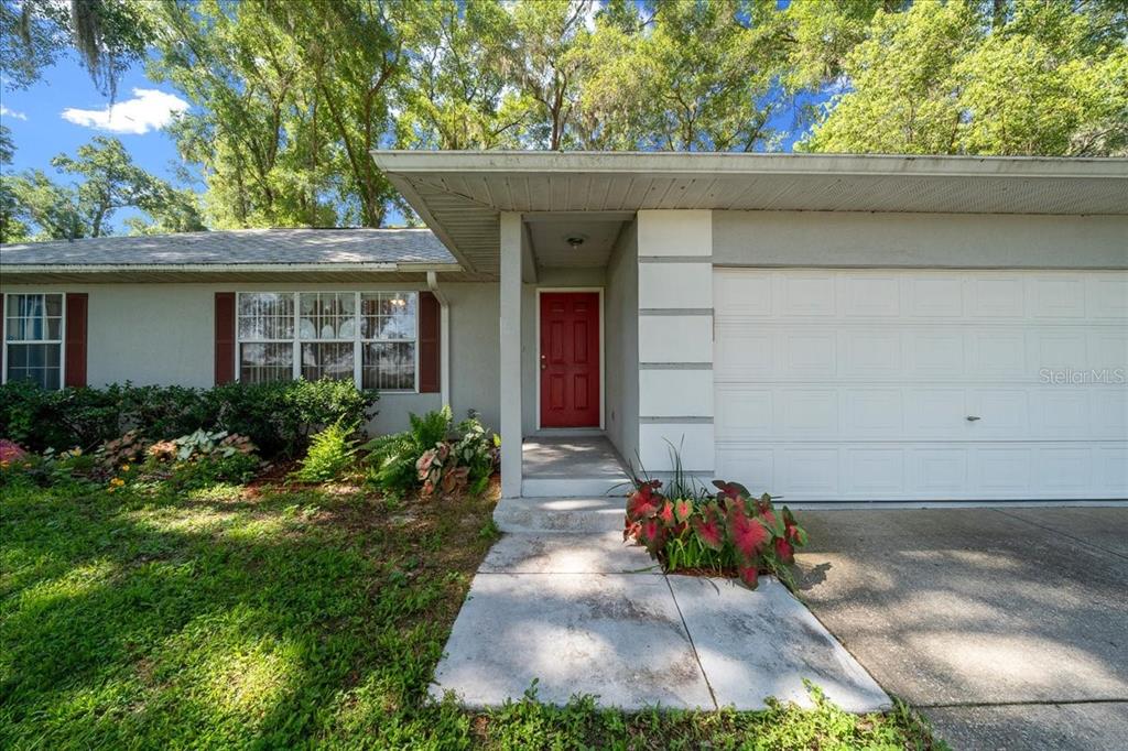 A single-story house features a red front door, a white garage door, and landscaped gardens — 3100 NE 35TH STREET, OCALA, FL