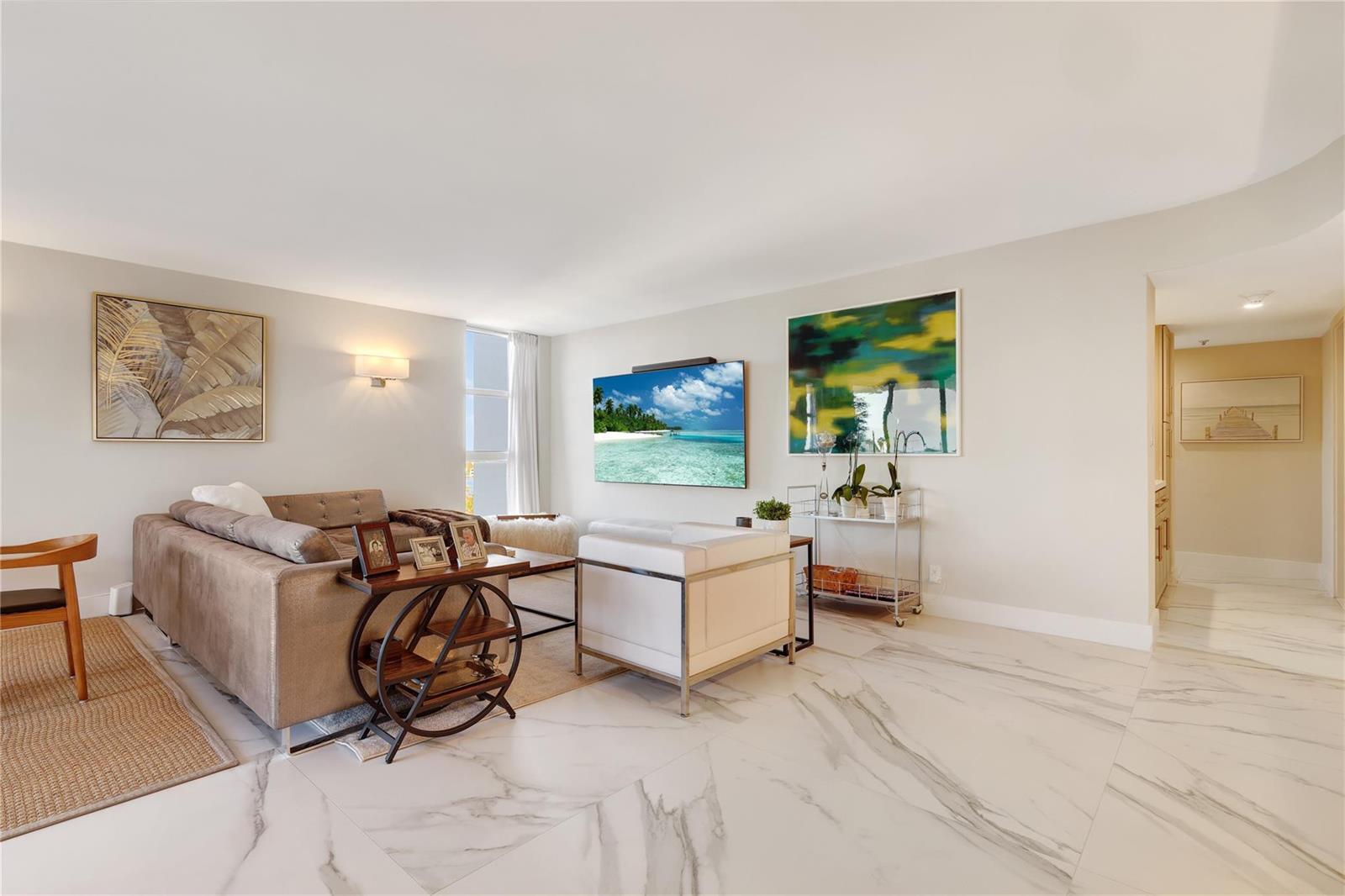 A modern living room features a sofa, white armchair, wall-mounted television, and marble tile flooring — 1361 S Ocean Boulevard # 910, Pompano Beach FL 33062, POMPANO BEACH, FL