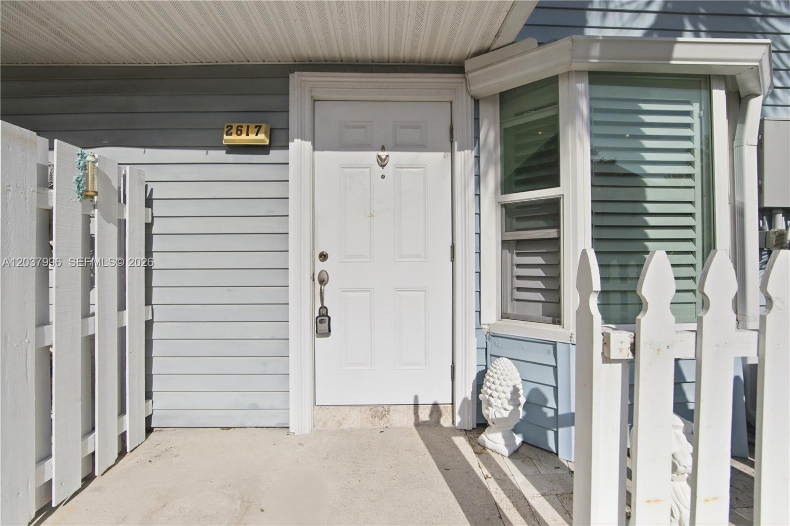 A white front door with a lockbox sits beside a bay window and fence — POMPANO BEACH, FL
