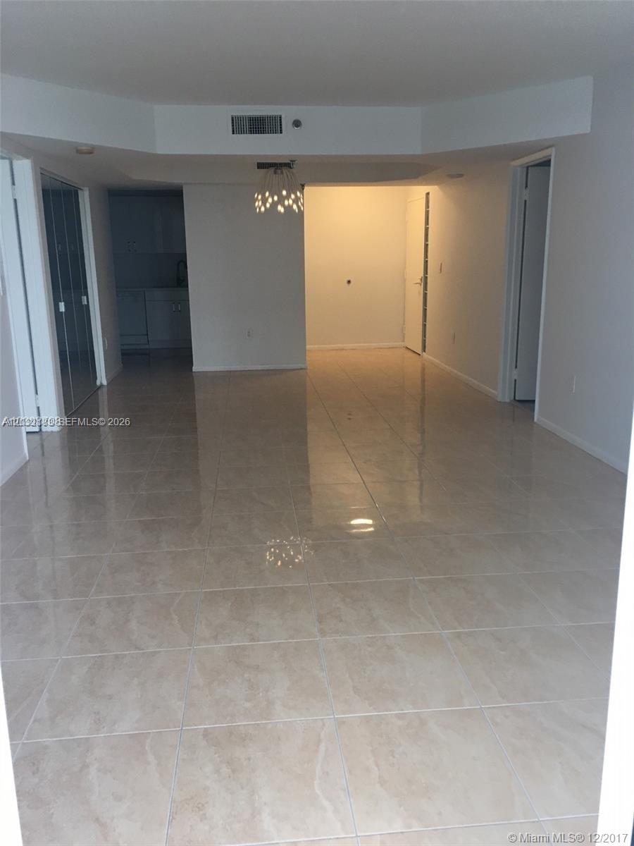 An empty room features light-colored tile flooring, a modern chandelier, and a recessed kitchen area — 9341 Collins Ave # 1006, Surfside FL 33154, SURFSIDE, FL