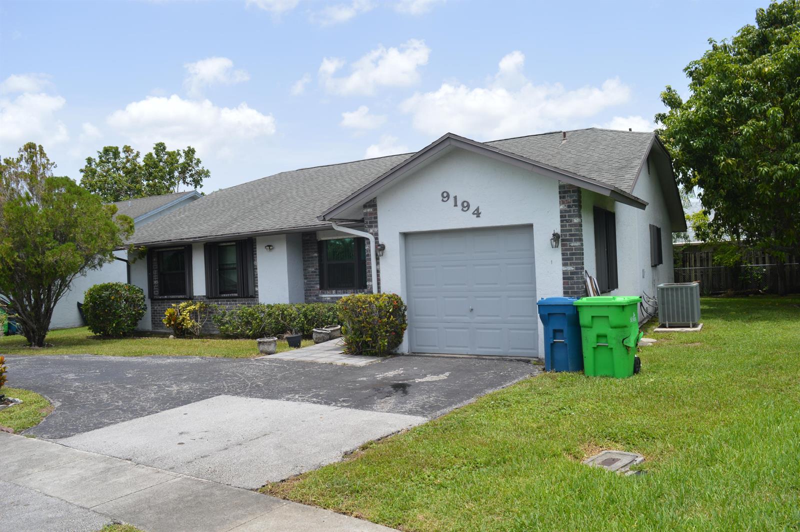 A single-story house with a gray garage door, brick accents, and two trash bins outside — 9194 NW 49TH Court, Sunrise FL 33351, SUNRISE, FL
