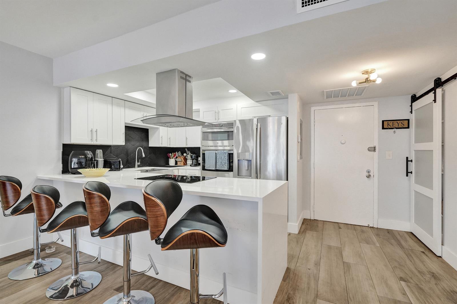 A modern kitchen features white cabinetry, stainless steel appliances, a breakfast bar, and wood-look flooring — 600 Three Islands Boulevard # 615, Hallandale Beach FL 33009, HALLANDALE BEACH, FL