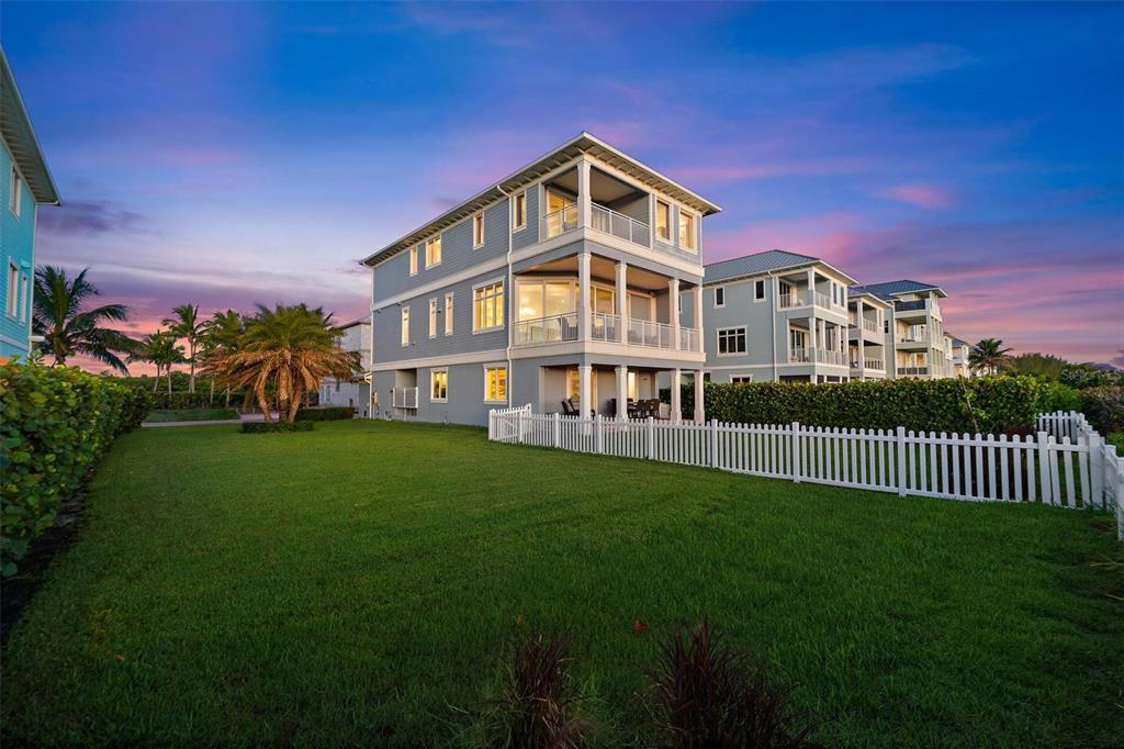 A three-story blue house with balconies stands behind a white picket fence at sunset — 4860 WATERSONG WAY, FORT PIERCE, FL