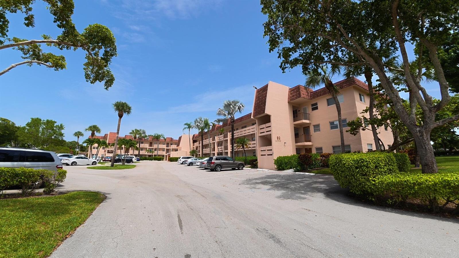 A multi-story apartment building with a parking lot and surrounding trees under a blue sky — 14790 Bonaire Boulevard # 210, Delray Beach FL 33446, DELRAY BEACH, FL