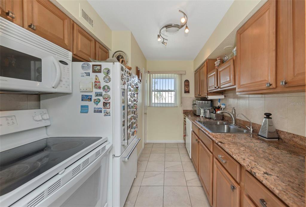 A galley kitchen features wood cabinets, granite countertops, white appliances, and light-colored tile flooring — 6976 SUNSET DRIVE S UNIT 6976, ST PETERSBURG, FL