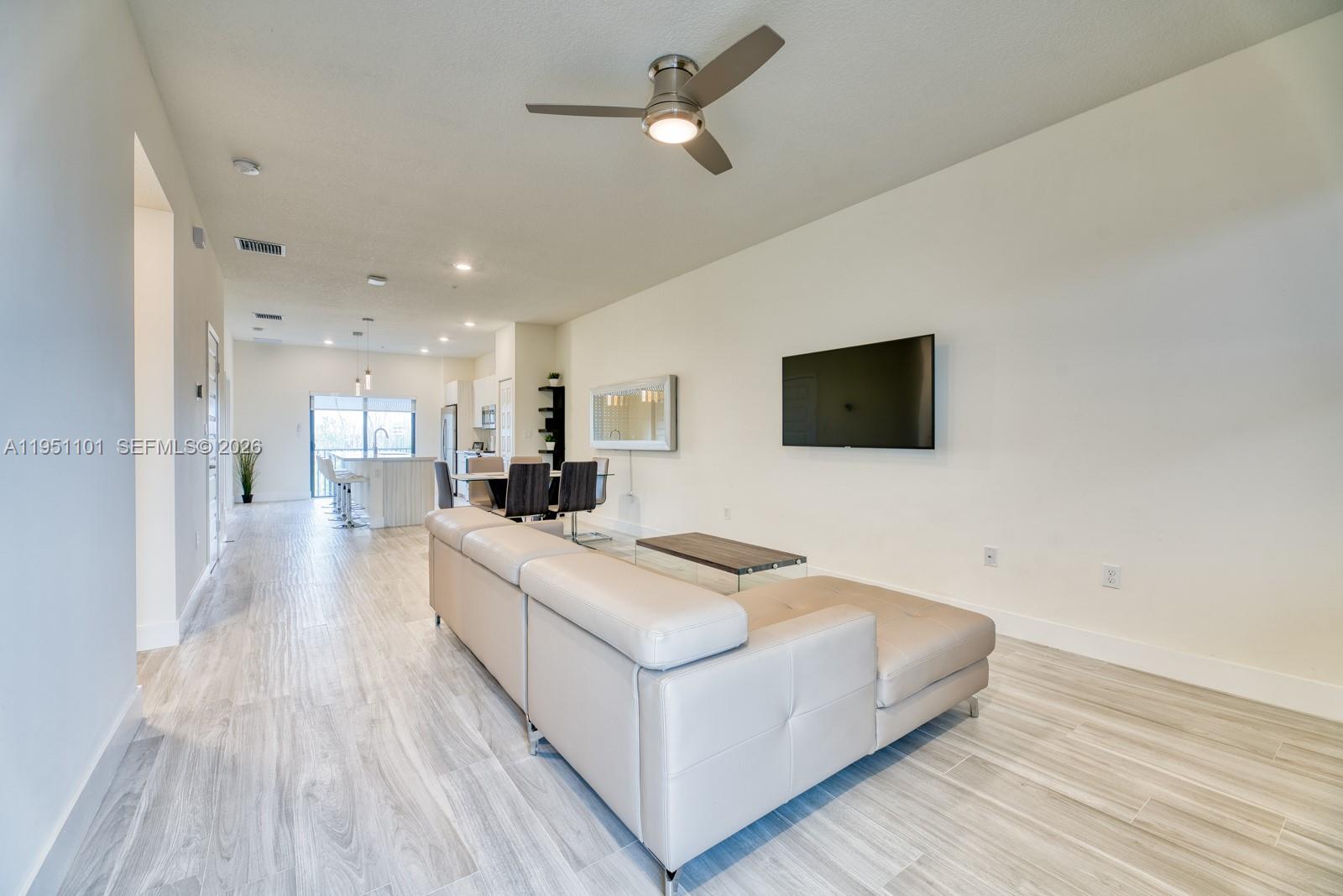 A modern living room features light wood-look tile flooring, a beige sectional sofa, and a wall-mounted television — 7831 NW 104th Ave # 23, Doral FL 33178, DORAL, FL