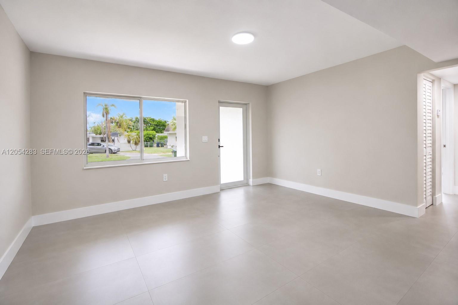 An empty room features light gray tile flooring, beige walls, a window, and a door — 6410 SW 17th St, West Miami FL 33155, WEST MIAMI, FL