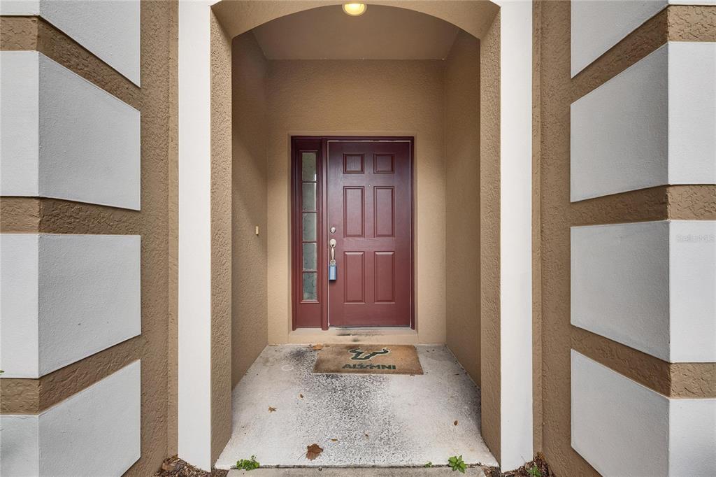 A covered entryway features a red front door with a sidelight and a concrete floor — 10328 CELTIC ASH DRIVE, RUSKIN, FL