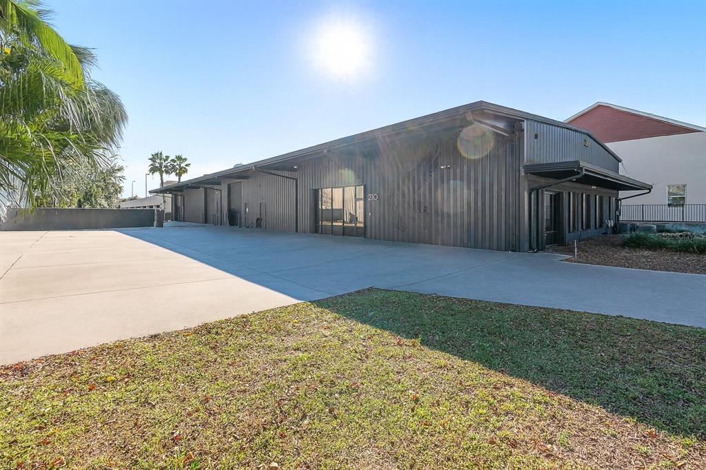 A low-rise industrial building with dark metal siding and a large concrete parking area — 210 E PALMETTO AVENUE, LONGWOOD, FL